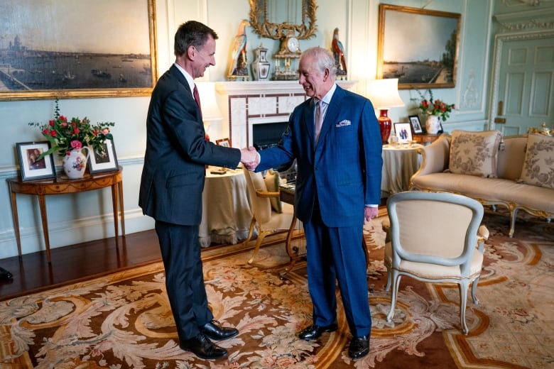 Two men in suits shake hands in an ornate parlour.