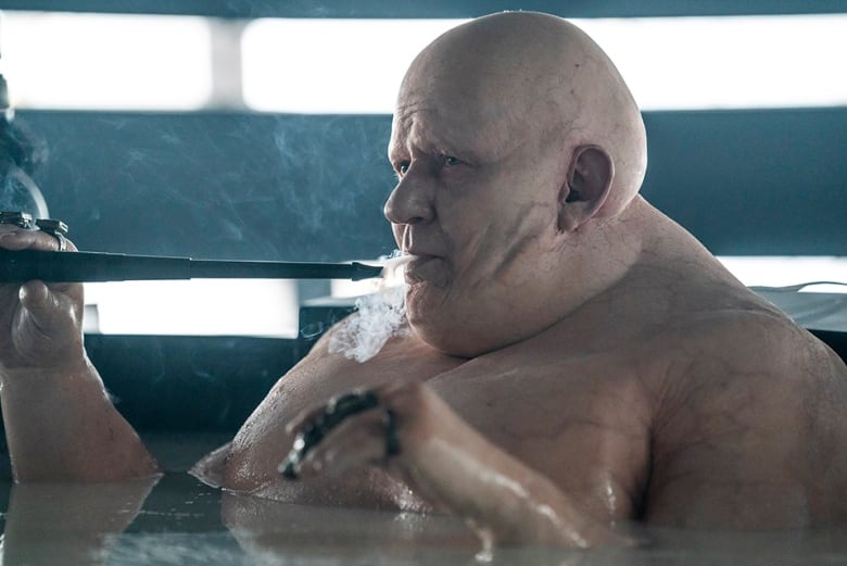 An incredibly large, bald man is bathing in a black tub. He is pale and blue veins can be seen under his skin. He is smoking from a long thin pipe. 