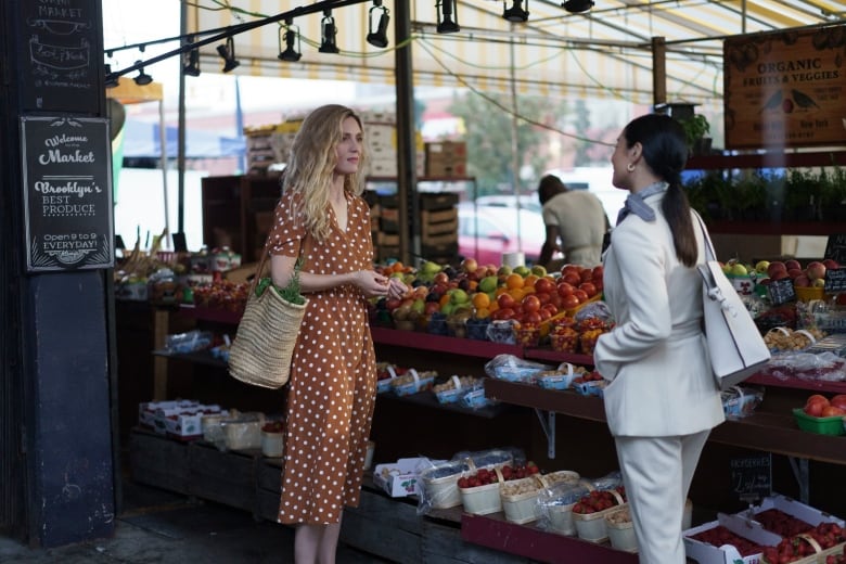 A blonde woman in a reddish brown dress with white polka dots is shown at a farmers market on the left. On the right is a brunette woman wearing a cream suit.