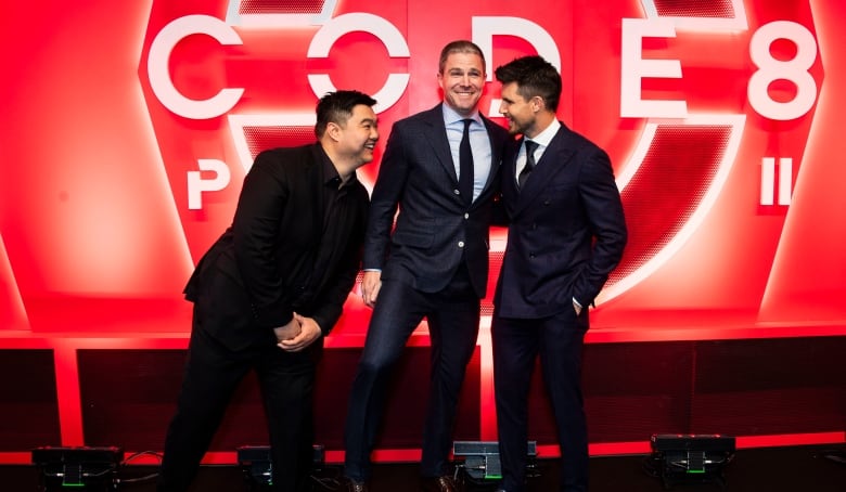 Three men in suits stand together on a red carpet. 