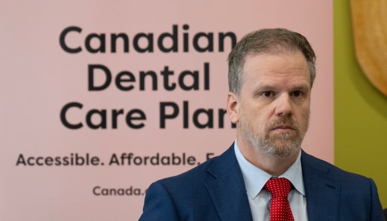 A man in a blue suit and red tie stands in front of a sign that reads "Dental Care Plan."