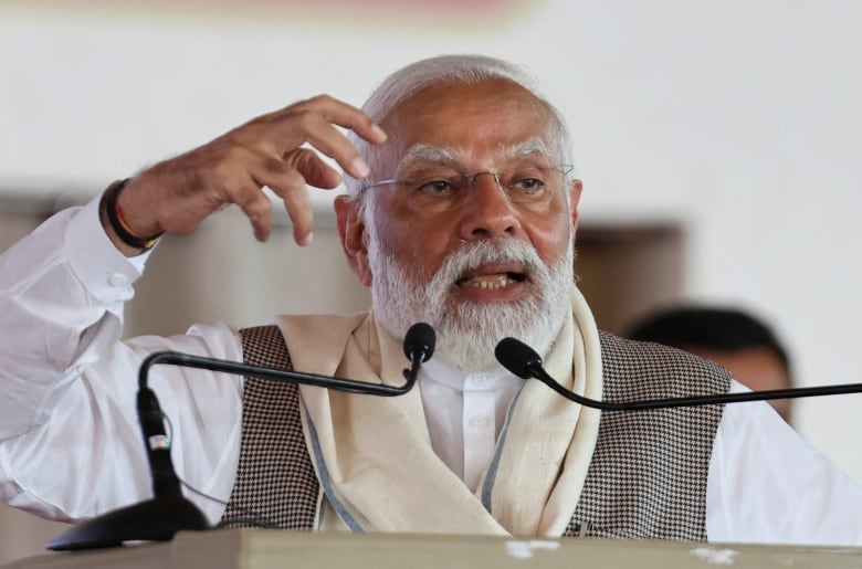 A man with white hair and a neat white beard gestures with both hands while speaking into a pair of microphones at a podium.