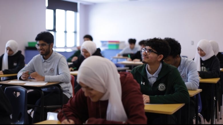 A young woman in a hijab puts her head over a desk, while other students in a classroom smile while looking up at a person off camera. 