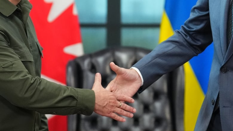 Two people shake hands in front of a red and white Canadian flag and a blue and yellow Ukrainian flag.