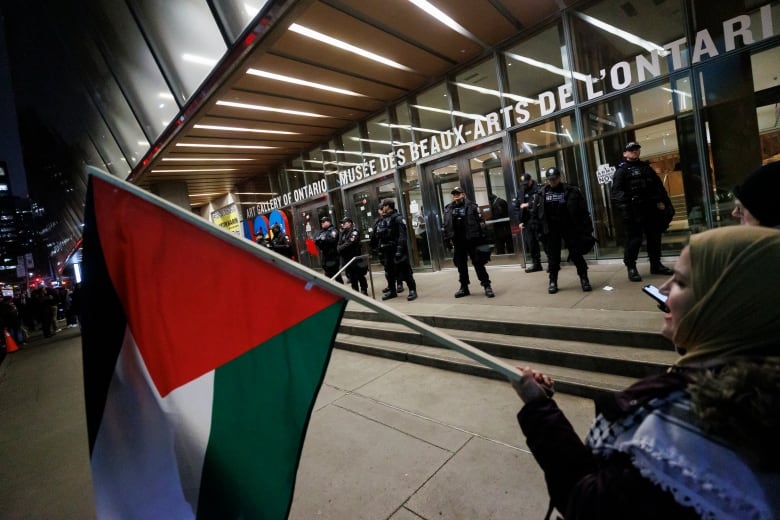 A woman waves a Palestinian flag outside of a building. A row of police officers stand in front of the entrance to the building.