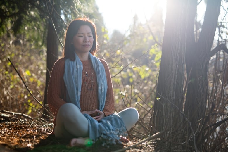 As Asian woman sits cross-legged in the forest with her hands in her lap and her eyes closed. 