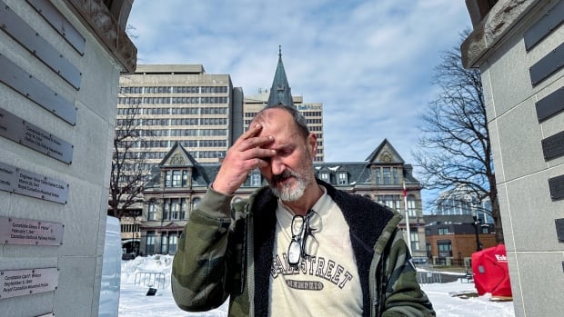 A man holds his head with his hand. He's standing in front of a large stone building in a snow-covered public square. 