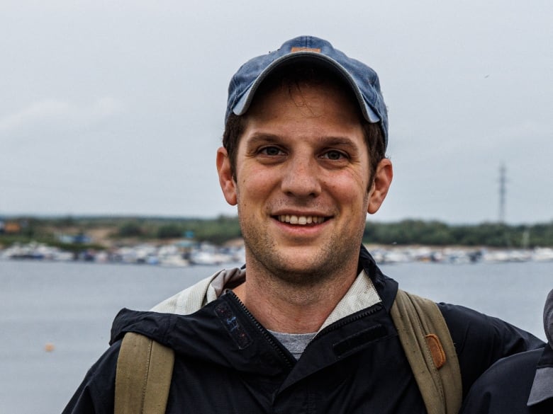 A man in a baseball cap and jacket with backpack straps is shown posing for a photo outdoors.