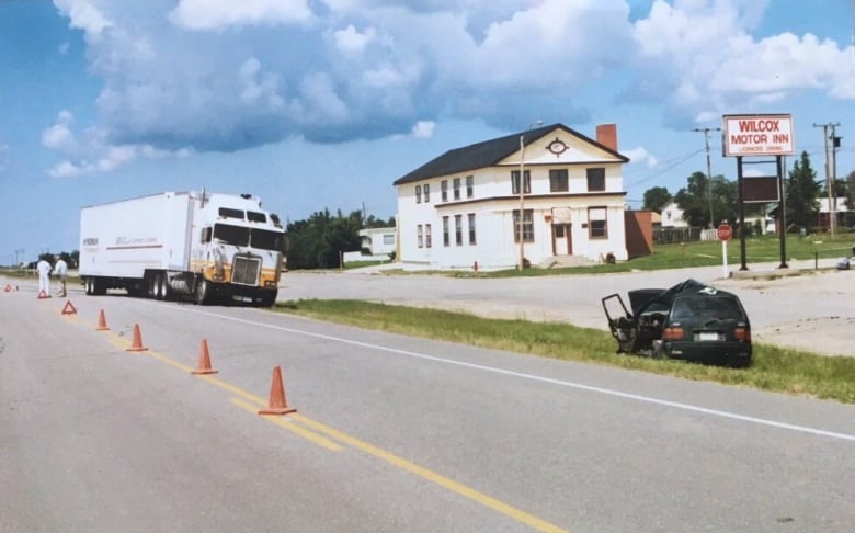 A mangled vehicle sits in the ditch of a highway. A bit farther down the road, a semi-trailer is pulled over to the side, facing it. Orange pilons bloack off the vehicles from traffic.