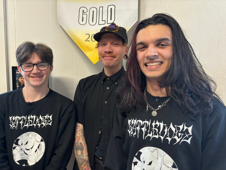 Three people wearing black stand in front of a banner that reads "gold."