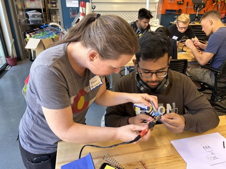 A woman stands next to a young man at a desk in a classroom, helping him use a soldiering iron. Behind them, a group of young people sit around a desk assembling devices.