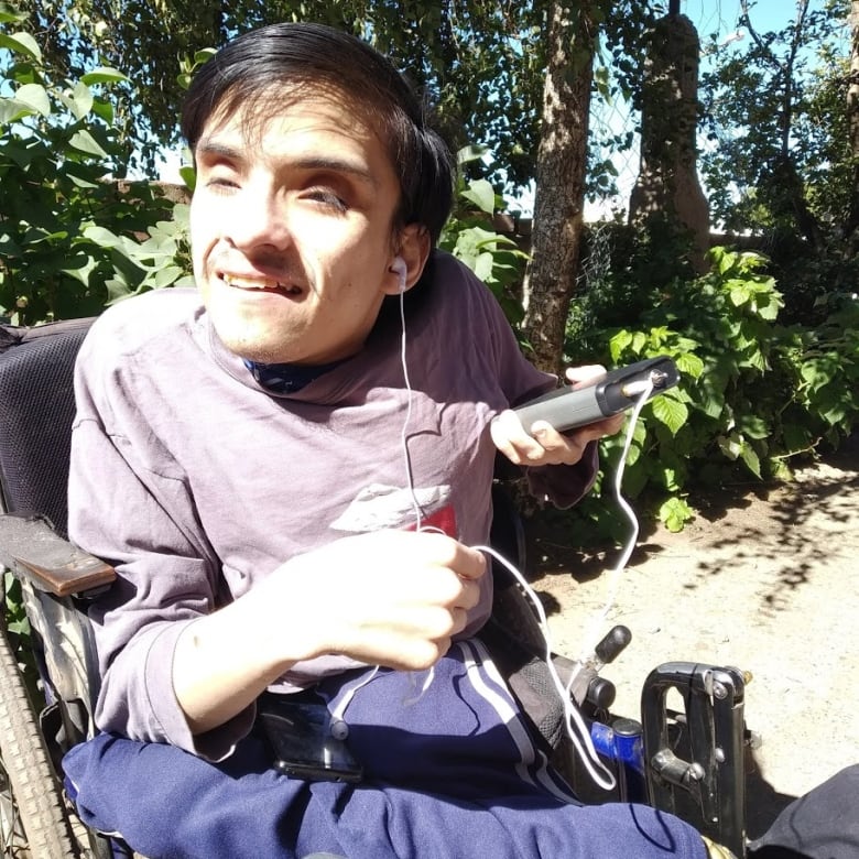 A man in a wheechair sits outside in the sun and listens to a small digital device through an earbud. 