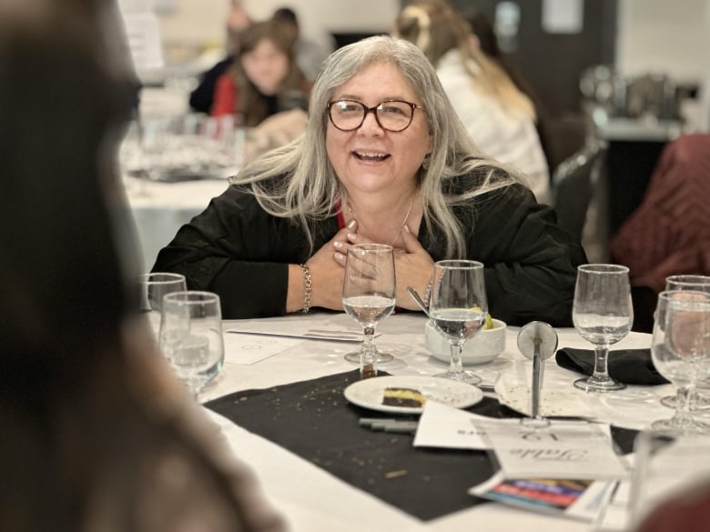 A bespectacled woman with long, white hair leans over a table in conversation.