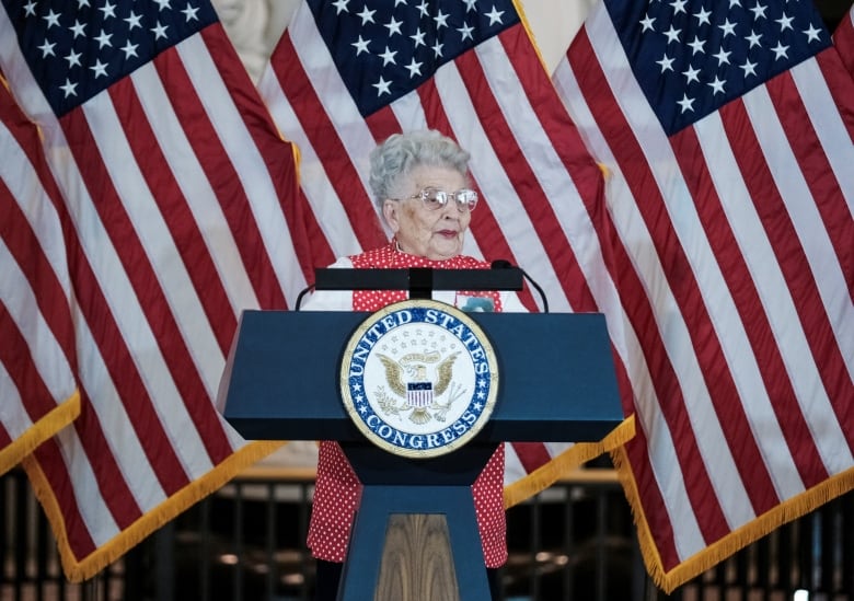 An elderly woman with white hair stands at a podium in front of three American flags, wearing a red and white polka-dot vest.