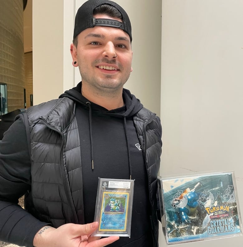 A man holds a Blastoise Pokemon card in one hand, and a box of unopened Pokemon cards in the other.