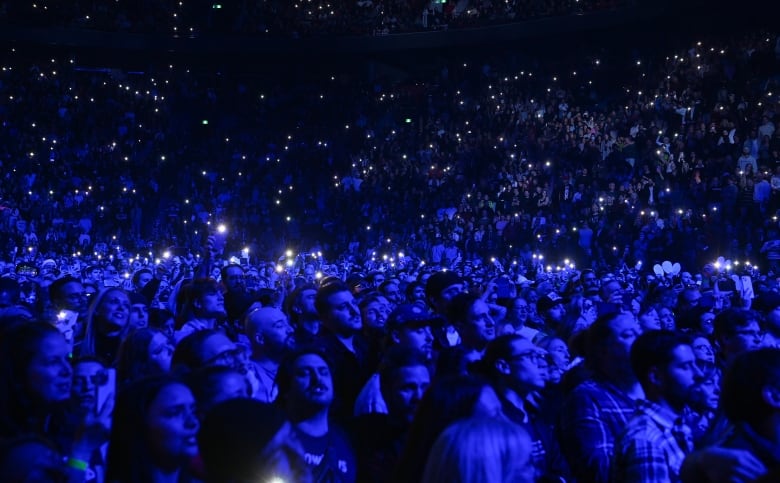 With some holding their cellphones up in the air, a crowd of people illuminated by blue light watch a stage, during a tribute to the late Karl Tremblay of Les Cowboys Fringants.