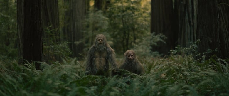 Two people in sasquatch costumes stand in the forest.