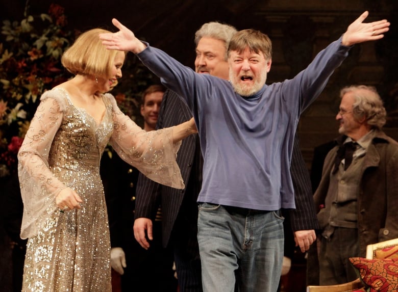 A bearded man is shown looking into the audience with arms spread and raised on a stage.