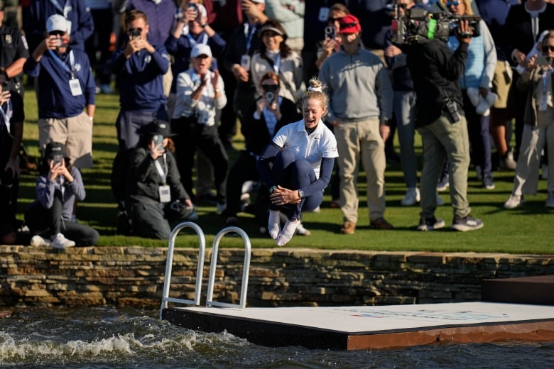 A woman in a white shirt and blue pants jumps into a lake after winning a golf tournament.