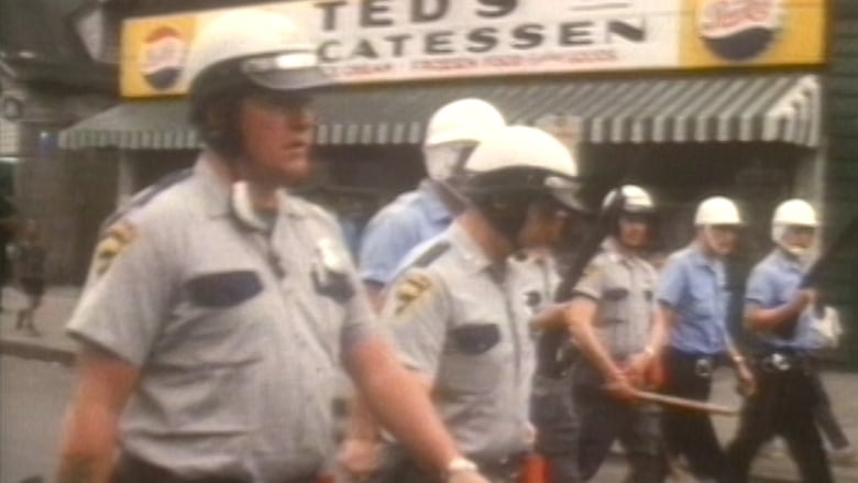 A blurry and old looking photo of police officers holding nightsticks and shotguns walking through an urban street is shown. 