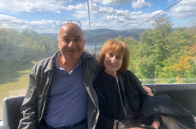 A man and a woman in their 60s sit on a gondala with a view of foliage behind them.