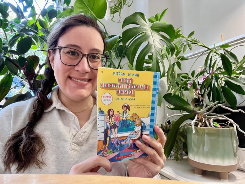 A woman with long hair in a braid holds up a book that says The Baby Sitters Club