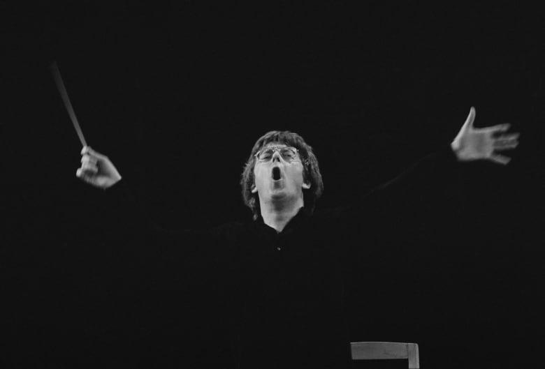 A musical conductor wearing glasses is shown with his mouth open in a black and white photograph.