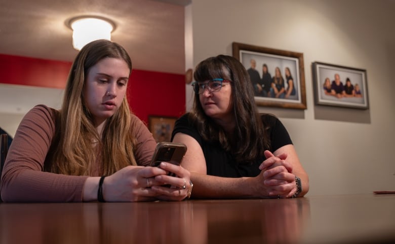 A woman with long blond hair sits at a dining table holding a smartphone as a woman with long dark hair and glasses sits next to her.