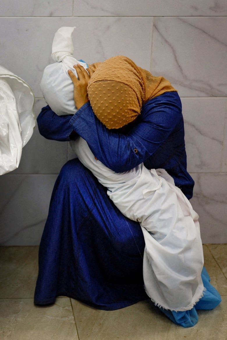 A woman in a blue dress and tan headscarf sits in a morgue in Khan Younis, Gaza, cradling the body of her niece that is wrapped in white cloth.