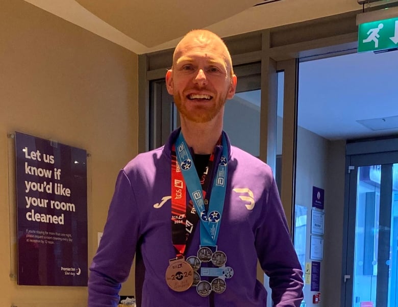 A smiling bald man with a short red beard stands in a hotel lobby with two medals around his neck, one of which has six circles.