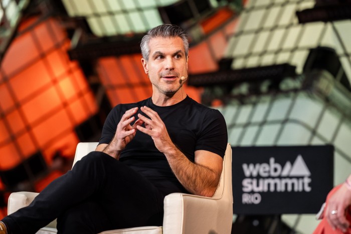 A man in black overalls on a sofa on a stage talking to someone to his left. Behind him is a sign that says ‘Web Summit Rio’