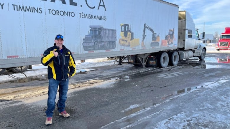 A man with a black ballcap, wearing a yellow and black coat and jeans is standing in front of an 18-wheeler truck.