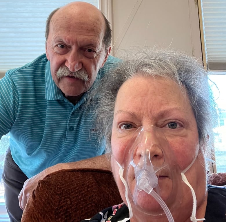 Old man with mustache sits next to his wife, an older woman with an oxygen mask.