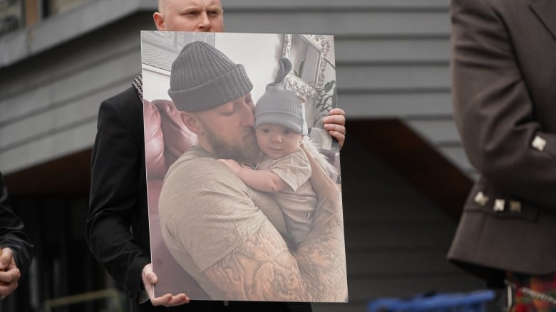 man holds a large picture of Jacob Flickinger in a beanie and t-shirt holding his infant son. 