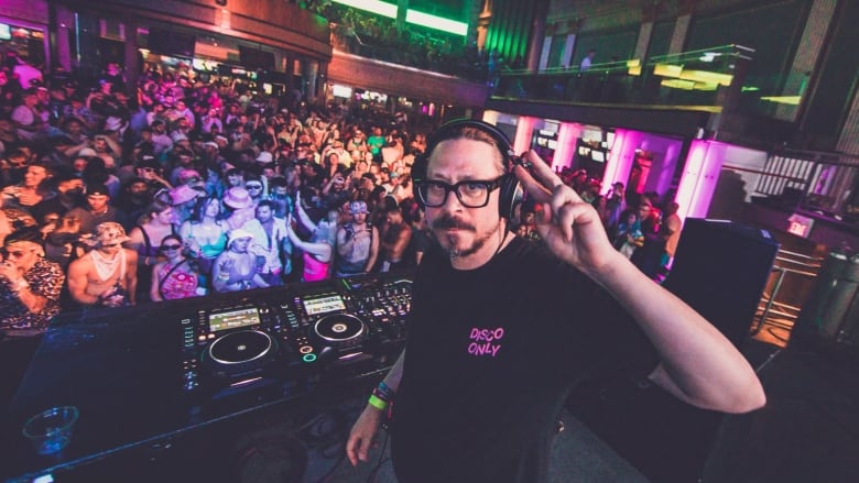 A man stands behind a DJ  booth with turntables, giving a peace sign to the camera. Behind him is a room with two levels filled with people dancing under bright lights.