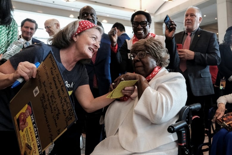 A woman in a wheelchair, with a red and white polka-dot scarf around her neck, surrounded by smiling people. 