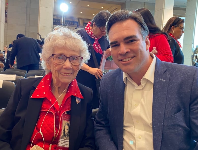 An elderly, white-haired woman in a bright red polka-dot blouse and black suit jacket sits in a crowded room next to a man in a suit. 