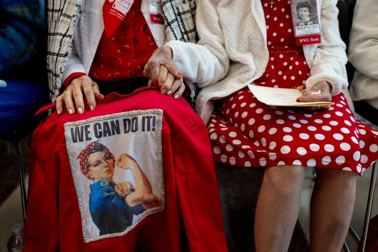 Two women in bright red and white polka-dots, pictured from the chest down, sit side-by-side holding hands. One has a bright red jacket draped over her knees with an image of the iconic wartime Rosie the River poster embroidered on the back. The poster shows a young woman in overalls with a red and white polka-dot scarf tied around her hair, flexing one arm, with the text: "We can do it!"