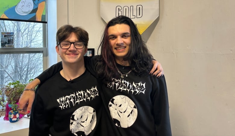 Two teens wearing black sweaters stand with their arms around each other's shoulders.