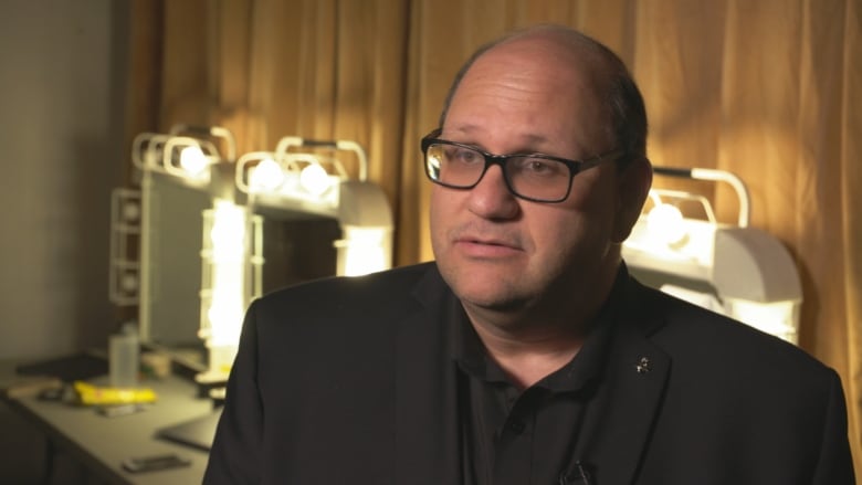 A man in glasses stands in front of dressing room mirrors