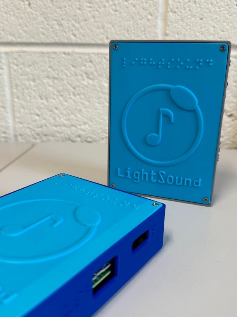 Two bright blue plastic rectangles, roughly the size of a Walkman, with USB ports on the side, and the word "LightSound" on the front in letters and braille along with a logo of a music note inside a circle.