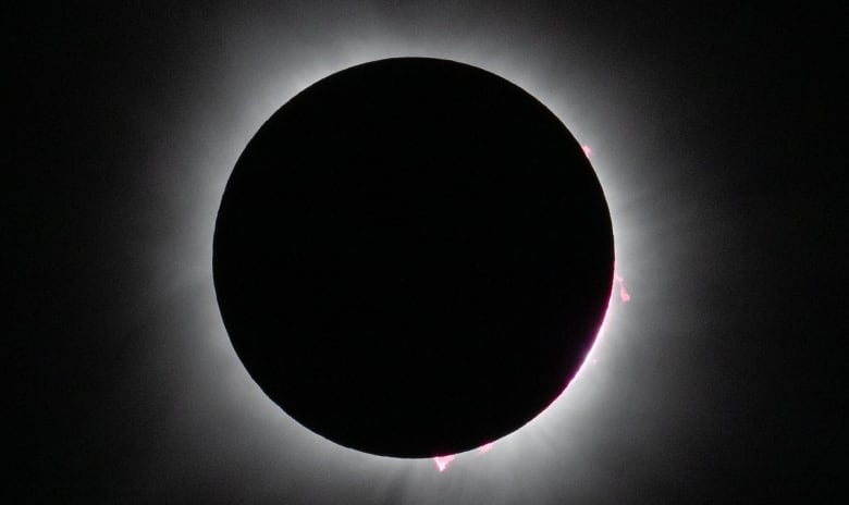 The silhouette of the moon, which appears black, completely covers the sun so that we can see detail in the sun's corona atmosphere as bright white light emanating from the sun's surface with tiny bits of pink protruding from the sun. 