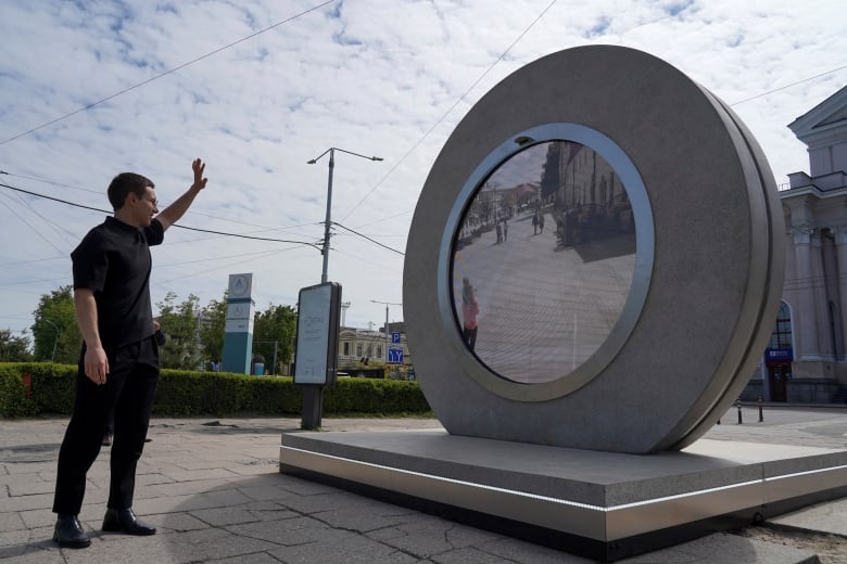 A man in black stands waving in front of a large metal circle with a screen showing a few people standing outside in the street.