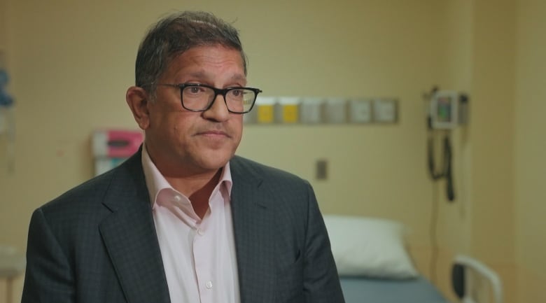 A man wearing a suit jacket and shirt with no tie in front of a hospital bed. 