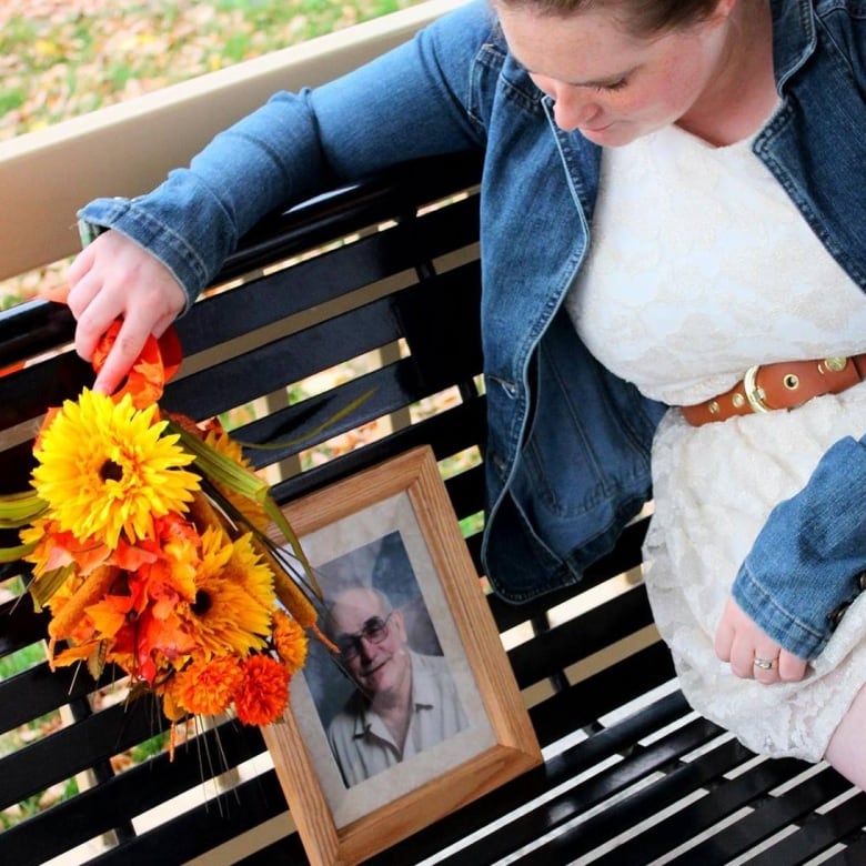 A woman sits on a park bench while holding a bouquet of flowers. She looks at a photo frame of a man.  