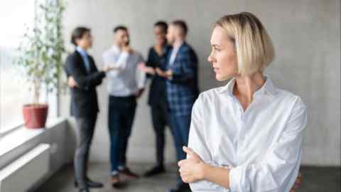 Male Coworkers Whispering Behind Back Of Unhappy Businesswoman