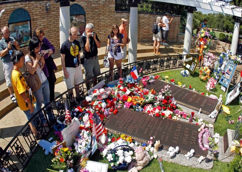 Several people stand, with some taking photographers, near memorial plaques outside on a property.