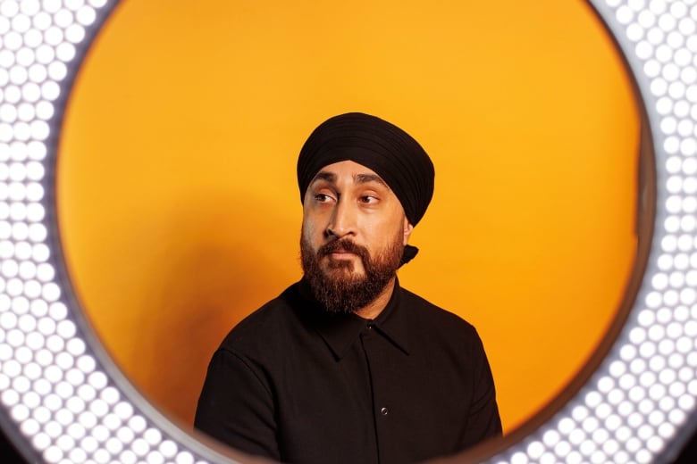 A man wearing a turban poses for a portrait in front of an orange backdrop with a ring light framing his face.