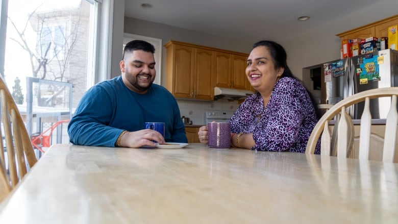 A young man and his mother sit at a kitchen table drinking tea and laughing. 