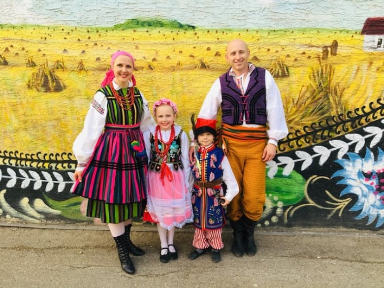 A family dressed in bright Polish costumes stand arm-in-arm, with the mom and daughter in pink skirts, a little boy in a blue outfit and a red hat, and the man at the far right wearing a vest and orange pants.